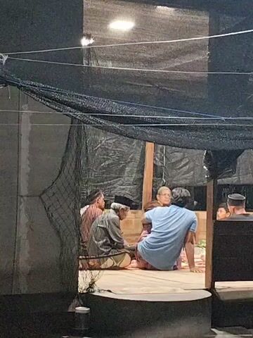 The video shows a group of people sitting on the floor, seemingly in a prayer or religious gathering. There are at least four individuals visible in the foreground, with more people possibly present in the background. The setting appears to be an outdoor or semi-outdoor space, possibly a prayer room or a sheltered area, as indicated by the netting and tarpaulin overhead. The lighting suggests it is either early morning or evening, or a dimly lit indoor space. The people are dressed modestly, with some wearing traditional Islamic attire like skullcaps. Their posture and the solemn atmosphere suggest a moment of reflection, prayer, or discussion related to religious matters. The overall mood seems calm and reverent. The location is identified as Gampongbaro, Indonesia.