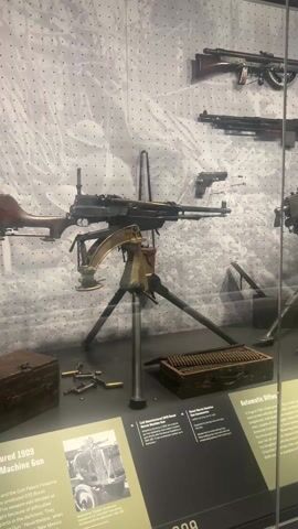 The video shows a museum exhibit case displaying a collection of historical firearms. The exhibit includes several machine guns mounted on tripods, along with other rifles and a handgun. Ammunition boxes and loose cartridges are also visible. Informational placards with text and photographs are positioned in front of the weapons. The background of the display features a large black-and-white image. The camera slowly pans from left to right, revealing the breadth of the exhibit. The room has a modern feel with visible ceiling lights. There is no visible human activity within the frame.
