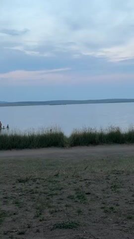 A video shows a body of water with a shoreline. On the left side of the screen, there are several people wading in the water. The sky above is cloudy with hues of blue and pink. The foreground consists of a dirt path with patches of grass.