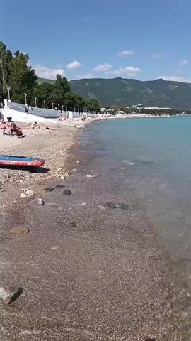 This is a video taken from a beach in Aderbiyevka, Russia. The scene opens with a view of the shoreline and the clear, turquoise water of the sea. The beach is made of pebbles and small stones, with some larger rocks scattered near the water's edge.

In the distance, a line of buildings stretches along the coast, backed by rolling green hills and mountains under a bright blue sky with a few scattered white clouds. The sun is shining, suggesting it is daytime, likely late morning or early afternoon.

There are several people visible on the beach, mostly spread out and appearing to be relaxing. Some are sitting or lying down, while others might be walking. A red object, possibly a deflated inflatable or a piece of equipment, is visible on the sand in the foreground. The water is calm, with gentle waves lapping at the shore. The overall impression is one of a peaceful and sunny day at the beach. There is no visible text in the video.
