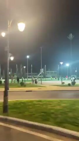 The video shows a nighttime scene of a well-lit city street alongside a park. Street lamps illuminate the road and surrounding areas. A white car is visible, initially moving and then parked on the side of the road. There are people in the background, appearing to be walking or standing in the park. The park has grassy areas, trees, and what appear to be tennis or badminton nets. A no-parking sign is also visible near the parked car. The sky is dark, and the overall atmosphere seems calm and peaceful.