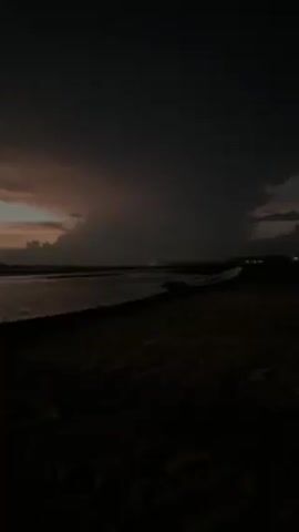 The video shows a dark, overcast scene near water. A landmass is visible in the foreground. In the distance, light reflects off the water, partially obscured by clouds. The sky is dark, with a hint of orange light breaking through the clouds on the left. The scene is mostly still and appears to be taken near twilight or at night.