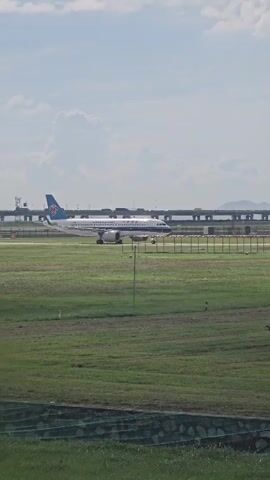 An airplane with a blue tail logo is on a runway. The plane is white with blue accents and the words China Southern appear on the side. The airplane appears to be moving slowly, potentially taxiing. In the background, there is a fence and a raised bridge or overpass. The ground is covered in green grass. The sky is partly cloudy.