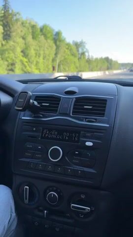 A person's hand with a ring on their ring finger is adjusting the volume on a car stereo. The car stereo has a digital display showing "04/08" and "ГРОМКОСТЬ 26". The dashboard of the car is visible, along with an air vent and a hazard light button. The car appears to be on a road with trees and a blue sky visible through the windshield.