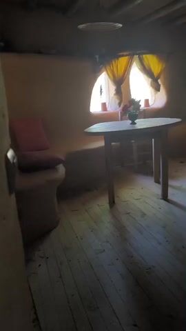 The video shows the interior of a rustic room with wooden floors and unfinished clay walls. A round wooden table with a vase of flowers is in the foreground, and a simple wooden chair is behind it. To the right, there is an arched fireplace opening with a metal grate inside. The walls have curved indentations and shelves. There is a bed with a mattress on a wooden frame, and a small white appliance on a bedside table. A wooden door is visible on the left wall. The room appears dimly lit, with light coming from a window with yellow curtains. The video pans to show a distinctive mushroom-shaped table with a television on top, and then pans across a bed. A close-up shot shows a window with a blue frame. The video concludes with a view of the curved clay walls and an exterior view of green foliage.