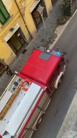 The video shows a street scene with several emergency vehicles and personnel. At the beginning, a red fire truck is partially visible, with a firefighter standing on the back. As the video progresses, more of the fire truck is revealed, along with a street and buildings. A firefighter is seen walking on the street. Later, a red car with "VIGILI DEL FUOCO" (Firefighters) written on it is shown, along with several firefighters and paramedics gathered around it. Two ambulances are also visible in the background. The scene appears to be an active emergency response situation.
