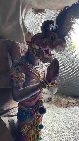 The video begins with a close-up of a statue of a creature with a colorful, serpent-like head and a human-like torso. The statue is wearing elaborate jewelry and has its hands pressed together in a gesture of prayer or greeting. The camera then pans to reveal a massive, dark-colored statue of a dragon or serpent, its body covered in intricate scales and designs. The dragon's tail is prominent, resting on rocks and gravel. The camera continues to move, showing more of the dragon's body winding through what appears to be an outdoor environment. The background shows a cloudy sky and some reddish, rocky structures. The video ends with a shot of the dragon's head against the sky, its eye visible and red. The overall impression is of a large, detailed sculpture, likely in a temple or religious site.