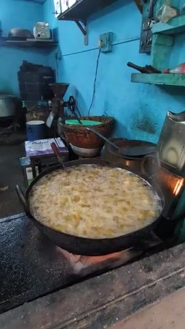 A close-up shot captures food being deep-fried in a large, black, wok-like pan placed on a stove. The pan is filled with bubbling oil, and numerous pieces of food are immersed and cooking. The stove's burners are visible underneath the pan, emitting flames. The background shows a rustic kitchen or food stall setting with blue walls, shelves holding various items like a grinder, baskets, and containers, and some equipment including a metal exhaust hood on the right. The overall impression is of a food preparation area where frying is taking place.