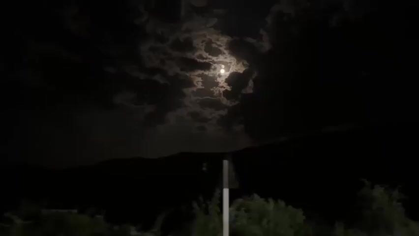 The video shows a night sky with the moon visible behind clouds. The landscape is dark with silhouettes of mountains and trees. There are also some lights visible in the distance on the mountain. The camera appears to be moving, possibly from a vehicle.