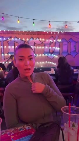 The video shows two women at an outdoor bar or restaurant, likely in the evening, given the lighting. The first woman, wearing a dark green top, is initially talking and gesturing with a smile. She has dark hair pulled back in a ponytail. The second woman, standing next to her, is wearing a dark brown jacket and camouflage pants. She has lighter hair, also in a ponytail. Both women are looking towards something or someone off-camera. In front of them on the table are menus and a plastic cup with a drink and red straws. The background features a colorful mural and string lights. Other people are visible at tables in the background, suggesting a bustling atmosphere.