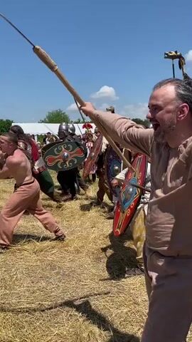 A group of men in historical clothing are engaged in a mock battle in a field. One man, shirtless and wearing light brown pants, is wielding a long stick. He is facing off against a group of men wearing helmets and holding shields and swords. Some of the shields are red and yellow, while others are green with a circular design. One person in a light brown tunic is standing with their back to the camera, observing the scene. In the background, there are white tents and a few spectators. The ground is covered in dry grass, and the sky is blue with some white clouds.