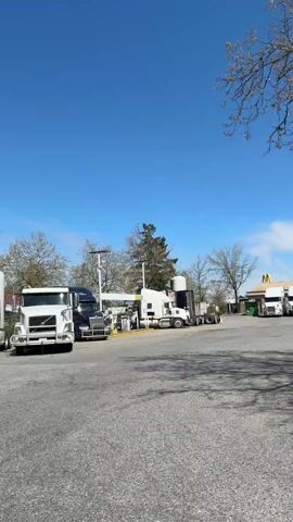 The video shows a truck stop under a clear blue sky with a few clouds. There are several large trucks parked around the gas pumps and a building with a "Flying J" sign. A McDonald's restaurant is also visible in the background. The camera pans from left to right, moving past some trees and bushes on a grassy area next to the parking lot. The camera focuses on the gas station and trucks as it pans.