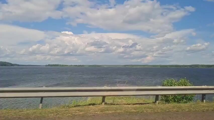 The video shows a wide expanse of dark blue water, likely a lake or large river, under a bright blue sky filled with fluffy white clouds. The far shore is lined with dark green trees. In the foreground, a metal guardrail runs horizontally across the bottom of the frame, with dry grass and some green vegetation visible beneath it. The camera appears to be moving, possibly from a vehicle, as indicated by the slight motion blur and the perspective of the guardrail. There are no people or specific objects of interest visible in the frame, other than the natural landscape and the man-made guardrail.
