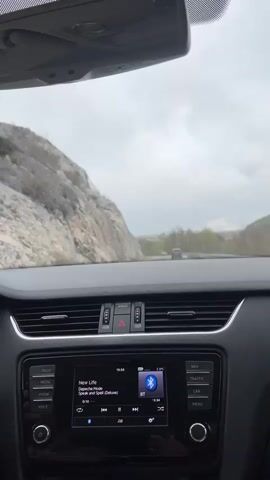 The image captures the view from the interior of a car, looking out through the windshield onto a road. The dashboard and infotainment system are prominently featured in the foreground.

Through the windshield, a winding road is visible, flanked by a steep, rocky mountainside on the left. In the distance, trees line the road, and another vehicle, possibly red, can be faintly seen. The sky is overcast and cloudy, indicating either a gray day or an impending change in weather. This landscape, with its rocky terrain, is consistent with the provided location context of Provalija, Montenegro.

Inside the car, the black dashboard occupies the lower half of the frame, featuring air vents with silver accents and a central hazard light button. The infotainment screen, illuminated, displays a time of 15:56. It shows that music is playing via Bluetooth (indicated by the "BT" icon). The current song is "New Life" by Depeche Mode, from the album "Speak and Spell (Deluxe)". Playback controls (play/pause, skip forward/backward) are visible, along with the song's elapsed time (0:13) and remaining time (-3:31). Around the screen, physical buttons are labeled "RADIO," "MEDIA," "VOICE" on the left, and "NAV," "TRAFFIC," "CAR," "MENU" on the right. A rearview mirror is partially visible at the top of the frame.

The scene suggests a car journey in the afternoon, with music playing, against a backdrop of a natural, rugged landscape. No people are visible, but the perspective implies a driver's or front passenger's view.