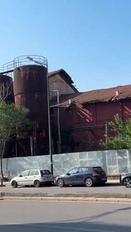The video shows a row of large, rusty industrial silos and a brick building with a red tile roof. Several cars are parked along the side of the road in front of the silos and building. The sky is clear and blue.