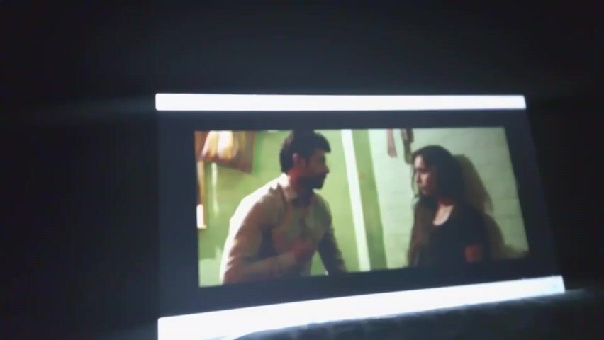 The video shows two people, a man and a woman, on a screen. The man is wearing a light-colored shirt and the woman is wearing a dark shirt. They appear to be in a room. The lighting is dim. The screen has white LED lights on the top and bottom.