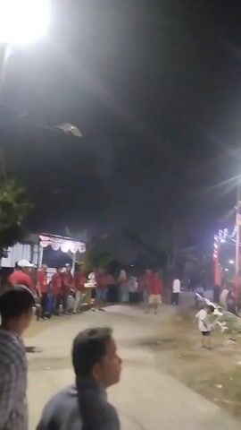 A nighttime outdoor gathering is taking place. The scene is illuminated by various lights, including string lights with colorful bulbs and spotlights. In the foreground, a man in a gray shirt is visible, looking to his left. The background shows a crowd of people sitting on mats on the ground, many of whom are wearing red shirts. There are also flags, banners, and what appear to be stalls set up along the sides of the gathering area. The ground is paved in some areas and appears to be dirt or sand in others, with some debris visible. There are also poles supporting the overhead decorations and lights. The overall impression is that of a community event or festival.