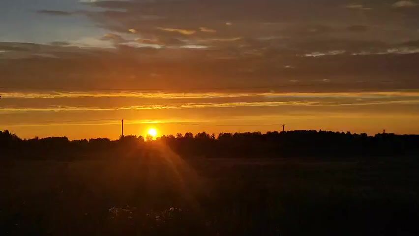 A wide shot captures a rural landscape at sunset. The sun, a bright orange orb, is partially obscured by a line of trees on the horizon, casting long rays of light across the sky. The sky is filled with dramatic clouds, painted in shades of orange, yellow, and gray. In the foreground, a dark field stretches out, with a few silhouetted plants visible. The scene is serene and peaceful, evoking a sense of natural beauty.