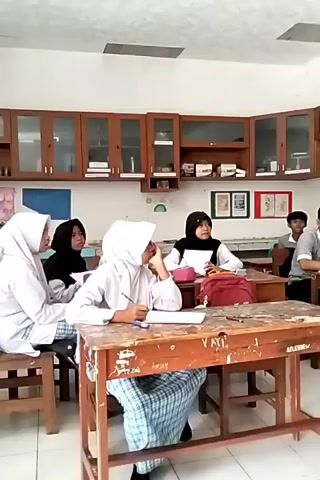 The video opens with a view of a classroom where several students are seated at wooden desks. A woman in a hijab, wearing a patterned dress, walks into the frame from the right, gesturing towards the students. She is partially out of focus as she moves. The camera then pans to show more students in the classroom. Some students are looking towards the front, others are looking down at their books or papers. The classroom has wooden cabinets along the walls filled with books and other items. There is a ceiling fan visible. The students appear to be engaged in a lesson or activity. The lighting suggests it is daytime.