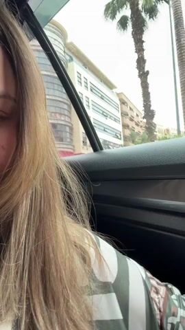 The video shows a view from inside a car driving on a city street. The car is stopped in traffic. Outside the window, there's a multi-story building with a curved, glass-covered corner and a pink store called "OBAYOLI" on the ground floor.  There are palm trees lining the street and other buildings visible in the background. Cars and a scooter pass by on the road.  The person filming is positioned behind the driver, partially showing the driver's blonde hair. The day appears overcast.