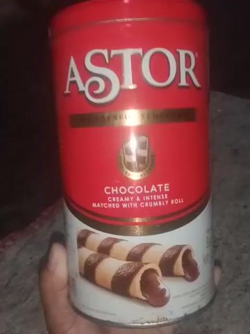 The video shows a person holding a cylindrical tin of Astor chocolate wafer rolls. The tin is red with white and gold lettering. The front of the tin features the Astor logo, an illustration of two chocolate wafer rolls, and text describing the product. The background is dark and appears to be a countertop or table. The person's hand is visible holding the tin, and their fingers are slightly curled around it. The lighting is dim, casting shadows on the tin and surrounding area. There are no other people or objects clearly visible in the frame.
