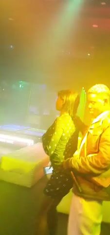 Two individuals are dancing in a dimly lit club or party environment. The person on the left, wearing a dark, possibly sequined outfit, is dancing with their back to the camera, their head tilted back. The person on the right, wearing a dark jacket and white pants, stands facing the first person, with their arms crossed. Various colored lights, including blue, purple, and green, illuminate the scene, creating a dynamic atmosphere. The background appears to show club furniture and lighting equipment, and there is some haze or fog in the air.