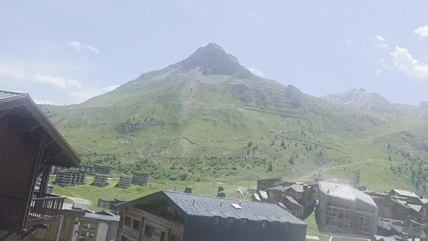 The video shows a panning view of a mountain village in a scenic alpine setting. In the foreground, there are several wooden and concrete buildings with pitched roofs. Some buildings appear to be multi-story apartments or hotels, with balconies and windows visible. A patch of vibrant red flowers in planters adds a touch of color. In the middle ground, more buildings of similar architectural style are clustered together, forming the heart of the village. Beyond the village, the landscape transitions to a lush green mountain slope dotted with trees. The upper part of the mountain is rocky and steep, with visible ski slopes or hiking trails carved into its face. The background is dominated by a majestic, snow-capped mountain range under a bright, partly cloudy sky. The overall impression is one of a tranquil mountain resort town, likely in the summer season given the green foliage. The camera is panning from left to right, revealing the expanse of the village and the surrounding mountainous terrain.