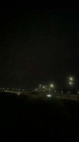 This media file captures a nighttime view of a highway or major road in Badhoevedorp, Netherlands. The scene is dominated by darkness, illuminated by the artificial glow of streetlights and vehicle headlights. The sky appears overcast and perhaps foggy, as there is a noticeable haze or mist that diffuses the light, creating a somewhat ethereal atmosphere.

The foreground is dark, suggesting a natural landscape like grass or scrub, with a fence or barrier visible along the edge of the roadway. In the mid-ground, the highway stretches into the distance, with lanes of traffic indicated by lines of lights. Several streetlights are positioned along the roadside, casting pools of yellow-white light onto the wet-looking road surface, hinting at recent rain or damp conditions. In the background, there are more distant lights suggesting urban development or other infrastructure.

The overall mood is quiet and somewhat isolated, typical of a late-night highway scene. The lack of visible people or distinct buildings places the focus on the infrastructure of the road and the atmospheric conditions. The time of day is clearly night, and the weather appears to be damp and possibly foggy. There is no visible text in the media file.