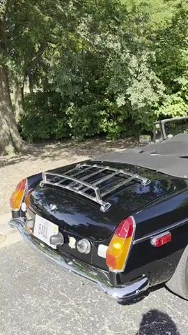 The video pans across the rear of a black convertible sports car. The car has a chrome luggage rack on the trunk, chrome bumpers, and red and amber taillights. The top is down, and the interior is black. The car is parked outdoors on an asphalt surface, with lush green trees and foliage in the background. The sunlight casts shadows on the ground and the car.