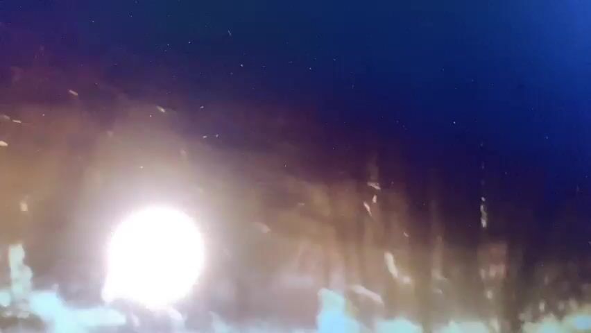 The video shows a bright, blurry light source in the lower left quadrant, possibly a sun or a strong artificial light. Streaks of light and indistinct shapes are visible in the background, suggesting motion or a hazy environment. The overall impression is of a low-resolution or out-of-focus image, with a dark blue hue dominating the upper portion of the frame. There are no discernible people, objects, or specific actions. The environment appears abstract and lacking in clear definition.