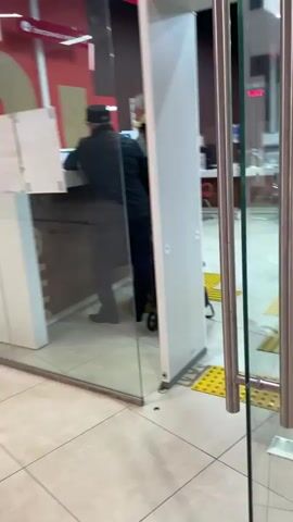 The camera pans across the interior of a post office or similar service center.  The space is brightly lit with fluorescent lighting and the floor is covered in off-white square tiles. There are designated pathways for the visually impaired, marked with textured yellow mats and bars on the floor.  Multiple service windows are visible, numbered from 01 to 06, each staffed with a worker seated in a red chair behind a counter.  Customers are at some of the windows. A small round table with a white top and base and a grey trash can are located in the foreground, toward the right of the frame.  People are engaged in transactions at the counters. The camera movement is a slow pan from left to right, providing an overview of the service area.