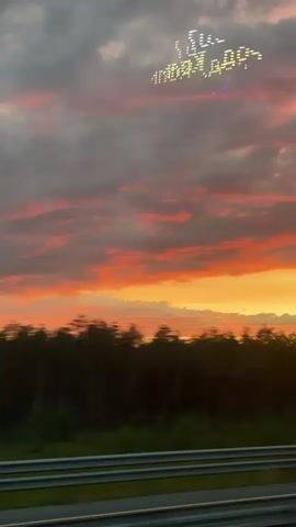 The video shows a sky at sunset with clouds in shades of orange, pink, and grey. In the upper portion of the sky, there is a formation of lights that appears to be spelling out a message. Below the sky is a dark line of trees, and below that, there are horizontal bars that resemble a guardrail or median barrier on a road. The image is slightly blurred, suggesting movement, possibly from a vehicle.