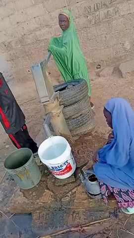 The scene shows a group of children around a water pump. Two girls, one wearing a green hijab and the other a blue hijab, are actively involved. The girl in green is operating the hand pump, drawing water into a white bucket labeled "BRIGHT PAINT". The girl in blue is kneeling down, using a smaller container to transfer water from the larger bucket. A young boy in a black and red tracksuit stands nearby, seemingly watching the activity.

Several containers are present, including the white bucket, a gray bucket, and the smaller container being used by the girl in blue. The pump is elevated by a stack of tires. The background features a brick wall and a dirt ground. The area around the pump is wet, suggesting recent use and spilled water.