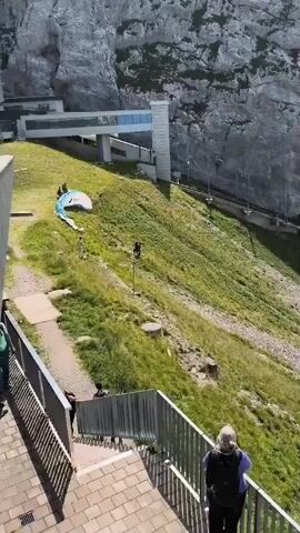 In a mountainous environment with a steep, grassy slope, a person wearing a blue and white paraglider is in the process of launching or landing. Several other individuals are visible on the slope, some appearing to assist with the paraglider and others observing. A modern building with large windows is situated at the top of the slope, and a metal railing with stairs is in the foreground where other observers are positioned. The weather appears to be clear and sunny.