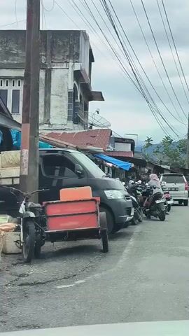 The video shows a street scene with several vehicles and buildings. On the left, a dark colored van and a red trailer are parked. Further down the street, motorcycles are parked along the side. A person in a headscarf is riding a motorcycle. Across the street, a truck is driving towards the camera. The street is paved and has a yellow line marking the lanes. The sky is cloudy. There are many electrical wires overhead. Buildings are visible on both sides of the street, with awnings and signs. In the background, mountains can be seen.