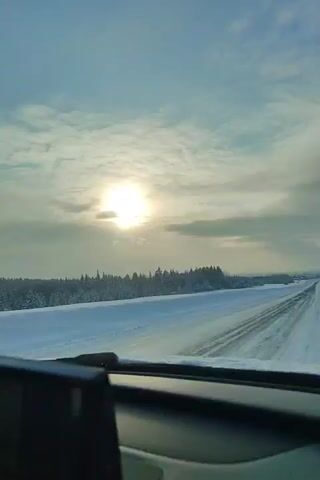The view from inside a car traveling down a snow covered road reveals a winter landscape. A forest lines the horizon, while the sky is partly cloudy with a bright sun and a rainbow-like phenomenon to the left of the sun. The car's dashboard and part of the roof are visible, framing the scene. Music is playing.