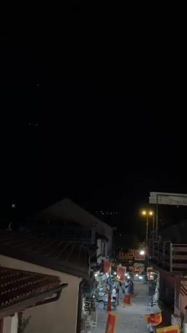 The video opens with a dark sky, with faint lights visible in the distance. As the camera pans down, a narrow street lined with buildings illuminated by streetlights becomes visible. Red flags with a yellow emblem hang from the buildings, indicating a festive or celebratory atmosphere. Several people are walking along the street, some appearing to be browsing at stalls. The architecture suggests an older, possibly European, town. The overall impression is of a lively street scene at night.