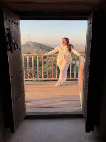The video shows a young woman standing on a wooden balcony. She is wearing a white tracksuit and a light-colored crossbody bag. She is leaning on the railing and looking at the camera. In the background, there is a landscape with palm trees, mountains, and a tower on a hilltop. The shot is framed by an open wooden door, adding depth to the scene. The sky is clear and the light is soft, suggesting it is either early morning or late afternoon. The woman appears relaxed and comfortable as she enjoys the view. The video appears to be taken by someone inside the building, looking out through the doorway.
