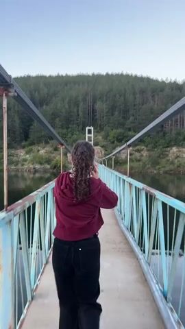 A person with curly brown hair tied back in a ponytail, wearing a maroon hooded sweatshirt and black pants, walks away from the camera on a suspension bridge. The bridge has light blue metal railings and a concrete walkway. The bridge spans over a calm body of water, likely a lake, with a forest covering a hill in the background. The sky is clear and blue. The camera pans to the right, revealing a wider view of the water and distant hills.