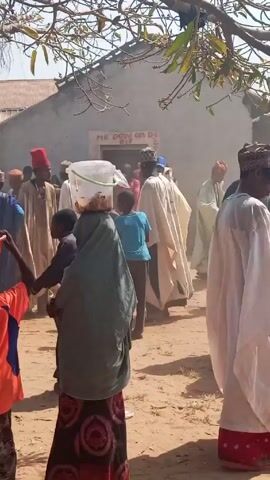 The video shows a group of people gathered outside a building. Many of the men are dressed in traditional robes and hats. Some individuals are wearing sunglasses. There are a few children present, one of whom is carrying an orange bowl, and another carrying a bucket on her head. The building has a corrugated metal roof, and the words "MR. DON ON DE 6IT" are written on it. A tree branch with sparse leaves hangs in the foreground. The ground appears to be dusty. Some of the people seem to be moving towards the building, while others are standing around, conversing. A loud musical tune plays in the background.