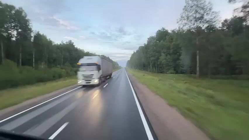 A dashcam view from a vehicle driving on a wet highway through a forest. The road has white lines marking the lanes and is surrounded by green grass and tall trees. The sky is overcast with some clouds. A large gray truck is visible passing on the left side of the frame at the beginning of the clip. The scene is dynamic, conveying a sense of motion and travel.