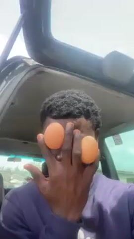A person stands in front of an open car trunk, obscuring their eyes with two brown eggs held in one hand. The person wears a navy blue sweatshirt and has short, dark, curly hair. Their fingernails appear to be painted a shade of orange. The car's interior is visible above the person's head, and a hazy outdoor scene can be seen in the background. The person appears to be looking directly at the camera.