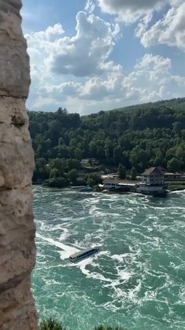 This media file showcases a breathtaking panoramic view of the Rhine Falls in Dachsen, Switzerland. The scene is dominated by the powerful Rhine River, its turquoise waters churning and frothing as it cascades over the falls. A white tour boat navigates through the turbulent water, appearing small against the immense scale of the falls.

The surrounding landscape is lush and verdant, with dense green forests covering the rolling hills that frame the river. In the distance, buildings, including two prominent modern high-rise towers, dot the landscape along the riverbanks and on the hillsides, suggesting a settled, populated area. Several smaller structures and what appear to be historical arched bridges are also visible along the river's edge, hinting at the rich history of the region.

The sky is a vibrant blue, adorned with fluffy white clouds, indicative of fair weather. The lighting suggests it is daytime, likely late morning or early afternoon, with the sun casting a bright, natural light on the scene. The perspective is from an elevated viewpoint, possibly a tower or a cliff edge, partially obscured by the rough, textured stone of a wall in the foreground, adding depth and a sense of exploration to the view. The overall mood is one of awe and natural beauty, with a touch of human presence integrated into the grand landscape. There is no visible text in the media file.