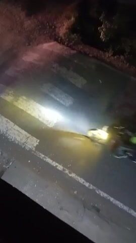 At night, a blurry video shows a road with a pedestrian crossing. A motorcycle with two people passes by, followed by another motorcycle with one person. The video is taken from an elevated position.