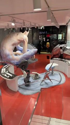 The video pans across a retail store with a vibrant red floor. In the center of the store, a large, abstract sculpture of a seated figure, colored in shades of blue and pink, dominates the space. Surrounding the figure are various display units showcasing metallic, abstract objects. A woman in a white top and dark pants is seen interacting with one of the displays near the back of the store. The store appears to be well-lit with overhead track lighting, and large windows offer a view of the street outside, where some signs are visible, including one that reads "ASIA L." The overall impression is a modern, artistic retail environment.