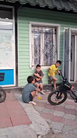 Four young boys are gathered outside a building. Two are standing, and two are sitting on the ground. One boy is sitting on the ground in the foreground, holding a yellow bottle. Another boy is sitting on a step behind him, with his arms crossed. A third boy is standing and leaning against the building, holding a drink. A fourth boy is standing further to the right. There are two electric scooters, one on the left and one on the right. The building has green siding and a white door with glass.