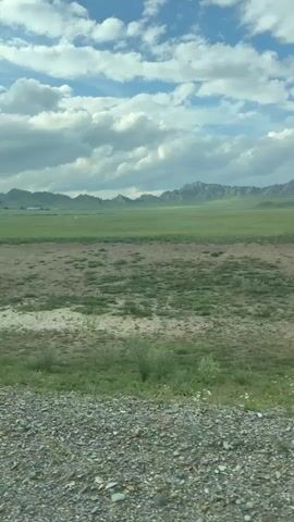 The video shows a landscape with a gravelly foreground, a dirt and grass middle ground, and a distant green field with mountains in the background under a cloudy sky. The camera appears to be moving, suggesting it is filmed from a vehicle. There are no people or discernible objects in the video besides the natural environment.