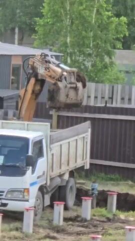 A construction scene shows an excavator dumping dirt into the bed of a white Nissan dump truck. The excavator's bucket is full of dirt and is positioned over the truck's bed. The truck is parked on a dirt and grass surface, and there are several concrete pilings with red caps in the foreground. In the background, there are corrugated metal buildings, a wooden fence, and trees. The scene appears to be a residential construction site.