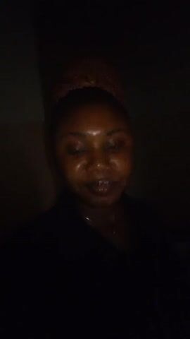 A woman is standing in a dark room. She is facing the camera and makes a peace sign with her fingers. Her hair is pulled back in a bun. She is wearing a dark-colored top and a thin necklace.