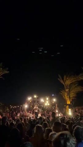 A crowded outdoor event at night. Palm trees are visible on the left and right sides of the frame. Many people are standing and gathered, facing a stage area where lights are illuminated and a person appears to be on the stage. String lights hang above the crowd and the stage area. The sky is dark, with some illuminated objects visible in the upper portion of the frame, and a bright light beam cuts through the scene at times.